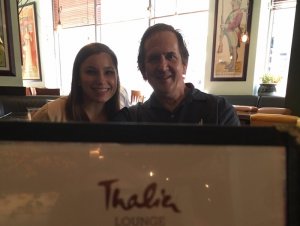 Elizabeth and David are in a cafe behind a sign that reads "Thalia Lounge"